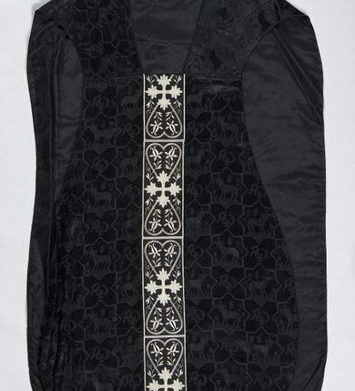 Zdjęcie nr 1: Ornat z czarnej tkaniny wiskozowej, żakardowej, z efektem adamaszku. Wzór o kompozycji sieciowej złożony z pól w kształcie gwiazdy ośmioramiennej (rombu opisanego na kwadracie) i czworoliści o płatkach zamkniętych łukiem dwuspadowym. W polach gwiaździstych przedstawienie Baranka w nimbie krzyżowym, wśród roślinności i z motywem chryzmonu. W kolejnych poziomych pasach Baranek zwrócony w prawo lub w lewo. W polu czteroliści splecione litery alfa i omega. 
Preteksty naszyte z tkaniny wiskozowej o splocie rządkowym, z haftem maszynowym nicią wiskozową, z motywem krzyży greckich o ramionach w kształcie liści, umieszczonych pomiędzy dwoma polami w kształcie serca, z wyrastającymi w górę i w dół parami fantazyjnych kwiatów na ulistnionych falistych łodyżkach. Na przecięciu ramion tylnej preteksty krzyżowej medalion w kształcie wydłużonego czworoliścia opisanego na kwadracie, ze stylizowanymi literami IHS z cienkim krzyżykiem łacińskim.
Podszewka z tkaniny syntetycznej o splocie rypsowym, w kolorze czarnym. Na podszewce metka z niebieskim napisem: Made by / THE MADONNA GUILDS / Especially For / Michigan Church Supply Co.