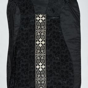 Zdjęcie nr 1: Ornat z czarnej tkaniny wiskozowej, żakardowej, z efektem adamaszku. Wzór o kompozycji sieciowej złożony z pól w kształcie gwiazdy ośmioramiennej (rombu opisanego na kwadracie) i czworoliści o płatkach zamkniętych łukiem dwuspadowym. W polach gwiaździstych przedstawienie Baranka w nimbie krzyżowym, wśród roślinności i z motywem chryzmonu. W kolejnych poziomych pasach Baranek zwrócony w prawo lub w lewo. W polu czteroliści splecione litery alfa i omega. 
Preteksty naszyte z tkaniny wiskozowej o splocie rządkowym, z haftem maszynowym nicią wiskozową, z motywem krzyży greckich o ramionach w kształcie liści, umieszczonych pomiędzy dwoma polami w kształcie serca, z wyrastającymi w górę i w dół parami fantazyjnych kwiatów na ulistnionych falistych łodyżkach. Na przecięciu ramion tylnej preteksty krzyżowej medalion w kształcie wydłużonego czworoliścia opisanego na kwadracie, ze stylizowanymi literami IHS z cienkim krzyżykiem łacińskim.
Podszewka z tkaniny syntetycznej o splocie rypsowym, w kolorze czarnym. Na podszewce metka z niebieskim napisem: Made by / THE MADONNA GUILDS / Especially For / Michigan Church Supply Co.