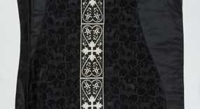 Zdjęcie nr 1: Ornat z czarnej tkaniny wiskozowej, żakardowej, z efektem adamaszku. Wzór o kompozycji sieciowej złożony z pól w kształcie gwiazdy ośmioramiennej (rombu opisanego na kwadracie) i czworoliści o płatkach zamkniętych łukiem dwuspadowym. W polach gwiaździstych przedstawienie Baranka w nimbie krzyżowym, wśród roślinności i z motywem chryzmonu. W kolejnych poziomych pasach Baranek zwrócony w prawo lub w lewo. W polu czteroliści splecione litery alfa i omega. 
Preteksty naszyte z tkaniny wiskozowej o splocie rządkowym, z haftem maszynowym nicią wiskozową, z motywem krzyży greckich o ramionach w kształcie liści, umieszczonych pomiędzy dwoma polami w kształcie serca, z wyrastającymi w górę i w dół parami fantazyjnych kwiatów na ulistnionych falistych łodyżkach. Na przecięciu ramion tylnej preteksty krzyżowej medalion w kształcie wydłużonego czworoliścia opisanego na kwadracie, ze stylizowanymi literami IHS z cienkim krzyżykiem łacińskim.
Podszewka z tkaniny syntetycznej o splocie rypsowym, w kolorze czarnym. Na podszewce metka z niebieskim napisem: Made by / THE MADONNA GUILDS / Especially For / Michigan Church Supply Co.