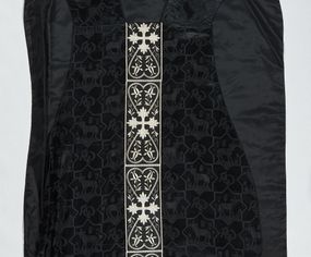 Zdjęcie nr 1: Ornat z czarnej tkaniny wiskozowej, żakardowej, z efektem adamaszku. Wzór o kompozycji sieciowej złożony z pól w kształcie gwiazdy ośmioramiennej (rombu opisanego na kwadracie) i czworoliści o płatkach zamkniętych łukiem dwuspadowym. W polach gwiaździstych przedstawienie Baranka w nimbie krzyżowym, wśród roślinności i z motywem chryzmonu. W kolejnych poziomych pasach Baranek zwrócony w prawo lub w lewo. W polu czteroliści splecione litery alfa i omega. 
Preteksty naszyte z tkaniny wiskozowej o splocie rządkowym, z haftem maszynowym nicią wiskozową, z motywem krzyży greckich o ramionach w kształcie liści, umieszczonych pomiędzy dwoma polami w kształcie serca, z wyrastającymi w górę i w dół parami fantazyjnych kwiatów na ulistnionych falistych łodyżkach. Na przecięciu ramion tylnej preteksty krzyżowej medalion w kształcie wydłużonego czworoliścia opisanego na kwadracie, ze stylizowanymi literami IHS z cienkim krzyżykiem łacińskim.
Podszewka z tkaniny syntetycznej o splocie rypsowym, w kolorze czarnym. Na podszewce metka z niebieskim napisem: Made by / THE MADONNA GUILDS / Especially For / Michigan Church Supply Co.