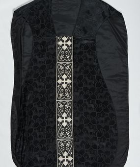 Zdjęcie nr 1: Ornat z czarnej tkaniny wiskozowej, żakardowej, z efektem adamaszku. Wzór o kompozycji sieciowej złożony z pól w kształcie gwiazdy ośmioramiennej (rombu opisanego na kwadracie) i czworoliści o płatkach zamkniętych łukiem dwuspadowym. W polach gwiaździstych przedstawienie Baranka w nimbie krzyżowym, wśród roślinności i z motywem chryzmonu. W kolejnych poziomych pasach Baranek zwrócony w prawo lub w lewo. W polu czteroliści splecione litery alfa i omega. 
Preteksty naszyte z tkaniny wiskozowej o splocie rządkowym, z haftem maszynowym nicią wiskozową, z motywem krzyży greckich o ramionach w kształcie liści, umieszczonych pomiędzy dwoma polami w kształcie serca, z wyrastającymi w górę i w dół parami fantazyjnych kwiatów na ulistnionych falistych łodyżkach. Na przecięciu ramion tylnej preteksty krzyżowej medalion w kształcie wydłużonego czworoliścia opisanego na kwadracie, ze stylizowanymi literami IHS z cienkim krzyżykiem łacińskim.
Podszewka z tkaniny syntetycznej o splocie rypsowym, w kolorze czarnym. Na podszewce metka z niebieskim napisem: Made by / THE MADONNA GUILDS / Especially For / Michigan Church Supply Co.