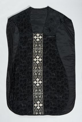 Zdjęcie nr 1: Ornat z czarnej tkaniny wiskozowej, żakardowej, z efektem adamaszku. Wzór o kompozycji sieciowej złożony z pól w kształcie gwiazdy ośmioramiennej (rombu opisanego na kwadracie) i czworoliści o płatkach zamkniętych łukiem dwuspadowym. W polach gwiaździstych przedstawienie Baranka w nimbie krzyżowym, wśród roślinności i z motywem chryzmonu. W kolejnych poziomych pasach Baranek zwrócony w prawo lub w lewo. W polu czteroliści splecione litery alfa i omega. 
Preteksty naszyte z tkaniny wiskozowej o splocie rządkowym, z haftem maszynowym nicią wiskozową, z motywem krzyży greckich o ramionach w kształcie liści, umieszczonych pomiędzy dwoma polami w kształcie serca, z wyrastającymi w górę i w dół parami fantazyjnych kwiatów na ulistnionych falistych łodyżkach. Na przecięciu ramion tylnej preteksty krzyżowej medalion w kształcie wydłużonego czworoliścia opisanego na kwadracie, ze stylizowanymi literami IHS z cienkim krzyżykiem łacińskim.
Podszewka z tkaniny syntetycznej o splocie rypsowym, w kolorze czarnym. Na podszewce metka z niebieskim napisem: Made by / THE MADONNA GUILDS / Especially For / Michigan Church Supply Co.