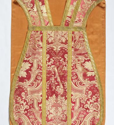 Zdjęcie nr 1: Ornat jednolity z czerwono-białej tkaniny półjedwabnej (?), żakardowej. Wzór o kompozycji sieciowej z motywem drobnych girland kwiatowych, rozłożystych centralnie umieszczonych układów roślinno-kwiatowych i elementów nawiązujących do ornamentu okuciowo-kartuszowego.

Galony tkane z nićmi o oplocie miedzianym złoconym w wątku, w dwóch szerokościach, z motywem czwórlistnych kwiatków i diagonalnych liści, brzegi z ząbkami.

Podszewka z tkaniny wiskozowej o splocie rządkowym, w kolorze pomarańczowym.