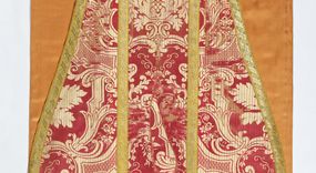 Zdjęcie nr 1: Ornat jednolity z czerwono-białej tkaniny półjedwabnej (?), żakardowej. Wzór o kompozycji sieciowej z motywem drobnych girland kwiatowych, rozłożystych centralnie umieszczonych układów roślinno-kwiatowych i elementów nawiązujących do ornamentu okuciowo-kartuszowego.

Galony tkane z nićmi o oplocie miedzianym złoconym w wątku, w dwóch szerokościach, z motywem czwórlistnych kwiatków i diagonalnych liści, brzegi z ząbkami.

Podszewka z tkaniny wiskozowej o splocie rządkowym, w kolorze pomarańczowym.