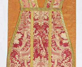 Zdjęcie nr 1: Ornat jednolity z czerwono-białej tkaniny półjedwabnej (?), żakardowej. Wzór o kompozycji sieciowej z motywem drobnych girland kwiatowych, rozłożystych centralnie umieszczonych układów roślinno-kwiatowych i elementów nawiązujących do ornamentu okuciowo-kartuszowego.

Galony tkane z nićmi o oplocie miedzianym złoconym w wątku, w dwóch szerokościach, z motywem czwórlistnych kwiatków i diagonalnych liści, brzegi z ząbkami.

Podszewka z tkaniny wiskozowej o splocie rządkowym, w kolorze pomarańczowym.