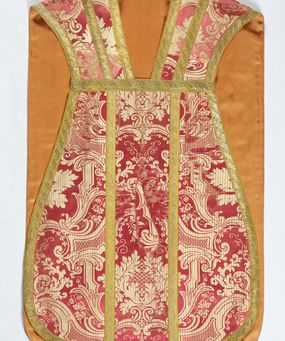 Zdjęcie nr 1: Ornat jednolity z czerwono-białej tkaniny półjedwabnej (?), żakardowej. Wzór o kompozycji sieciowej z motywem drobnych girland kwiatowych, rozłożystych centralnie umieszczonych układów roślinno-kwiatowych i elementów nawiązujących do ornamentu okuciowo-kartuszowego.

Galony tkane z nićmi o oplocie miedzianym złoconym w wątku, w dwóch szerokościach, z motywem czwórlistnych kwiatków i diagonalnych liści, brzegi z ząbkami.

Podszewka z tkaniny wiskozowej o splocie rządkowym, w kolorze pomarańczowym.