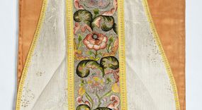 Zdjęcie nr 1: Boki ornatu z białej tkaniny wiskozowej o splocie rypsowym z malowanym wzorem z motywów okazałych bukietów drobnych kwiatów przewiązanych kokardą i mniejszych gałązek kwiatowych. W górnej części ornatu, na wysokości ramion, tkanina wiskozowa o tle w splocie rypsowym, żakardowa, z wzorem o kompozycji pasowej z ułożonych poziomo bukiecików drobnych kwiatków z listkami i luźno rozrzuconych małych kwiatków.

Preteksty w partii tła założone haftem kładzionym nicią o oplocie metalowym w kolorze srebrnym; wzór haftowany barwnymi nićmi ściegiem cieniowanym,  z motywem gałązek z fantazyjnie wygiętymi liśćmi i barwnymi kwiatami, m.in. róży i goździka. Dolna część tylnej preteksty uzupełniona podobnym haftem.

Galony tkane z nici sztucznych, trójbarwne (tło żółte, wzór biało-czerwony), w dwóch szerokościach i o dwóch wzorach: 1. przy pretekstach szerszy, z motywem białych krzyżyków greckich, motywem w kształcie litery X i rombów z wpisanym małym krzyżykiem, wszystkie z czerwonymi środkami; 2. na brzegach, z motywem białych krzyżyków greckich i ośmiopłatkowych kwiatków, z czerwonymi środkami i przedzielone czerwonymi kostkami. U dołu przedniej preteksty wstawiony fragment galonu tkanego z nićmi o oplocie miedzianym srebrzonym w wątku, z motywem falistej prążkowanej wstęgi i trójklapowych listków.

Podszewka z tkaniny wiskozowej o splocie rządkowym, w kolorze pomarańczowym.