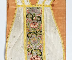 Zdjęcie nr 1: Boki ornatu z białej tkaniny wiskozowej o splocie rypsowym z malowanym wzorem z motywów okazałych bukietów drobnych kwiatów przewiązanych kokardą i mniejszych gałązek kwiatowych. W górnej części ornatu, na wysokości ramion, tkanina wiskozowa o tle w splocie rypsowym, żakardowa, z wzorem o kompozycji pasowej z ułożonych poziomo bukiecików drobnych kwiatków z listkami i luźno rozrzuconych małych kwiatków.

Preteksty w partii tła założone haftem kładzionym nicią o oplocie metalowym w kolorze srebrnym; wzór haftowany barwnymi nićmi ściegiem cieniowanym,  z motywem gałązek z fantazyjnie wygiętymi liśćmi i barwnymi kwiatami, m.in. róży i goździka. Dolna część tylnej preteksty uzupełniona podobnym haftem.

Galony tkane z nici sztucznych, trójbarwne (tło żółte, wzór biało-czerwony), w dwóch szerokościach i o dwóch wzorach: 1. przy pretekstach szerszy, z motywem białych krzyżyków greckich, motywem w kształcie litery X i rombów z wpisanym małym krzyżykiem, wszystkie z czerwonymi środkami; 2. na brzegach, z motywem białych krzyżyków greckich i ośmiopłatkowych kwiatków, z czerwonymi środkami i przedzielone czerwonymi kostkami. U dołu przedniej preteksty wstawiony fragment galonu tkanego z nićmi o oplocie miedzianym srebrzonym w wątku, z motywem falistej prążkowanej wstęgi i trójklapowych listków.

Podszewka z tkaniny wiskozowej o splocie rządkowym, w kolorze pomarańczowym.